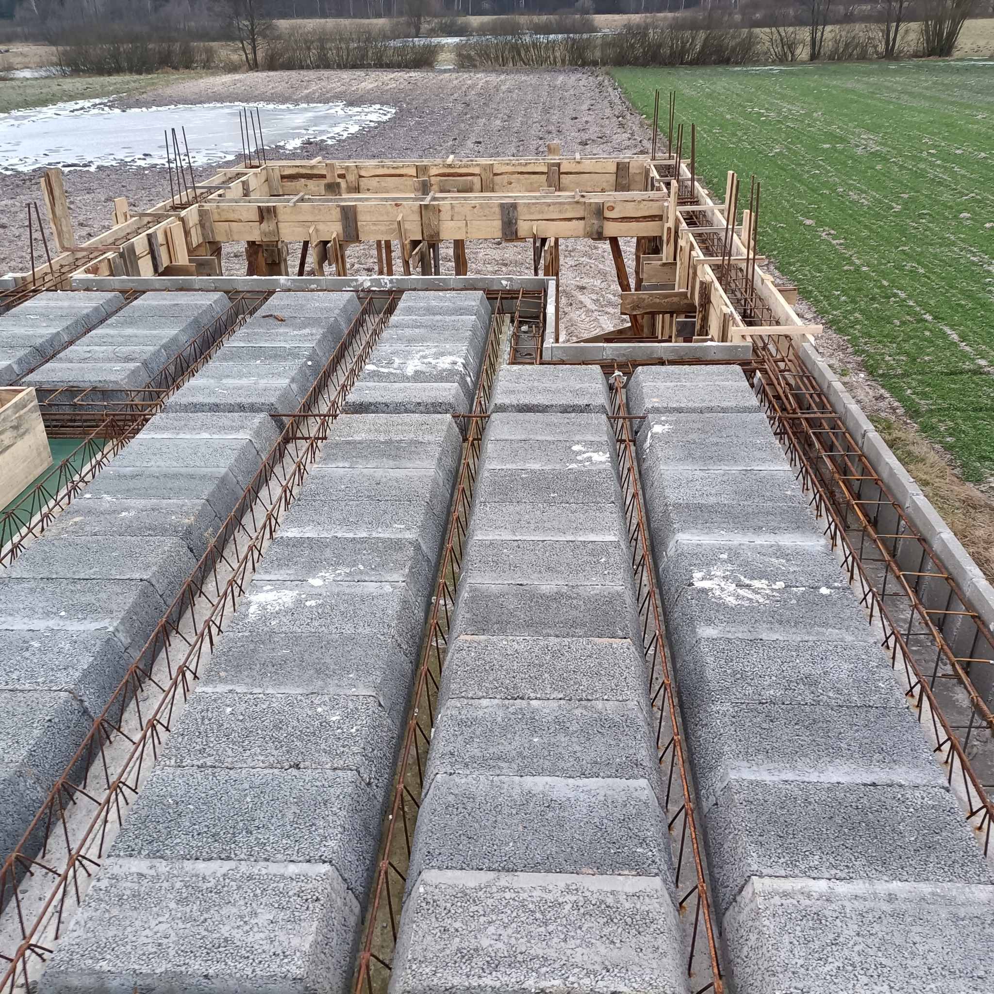Konstrukcja stropu Teriva z pustaków betonowych i zbrojenia stalowego, widok z góry na placu budowy, w tle pole i resztki śniegu.