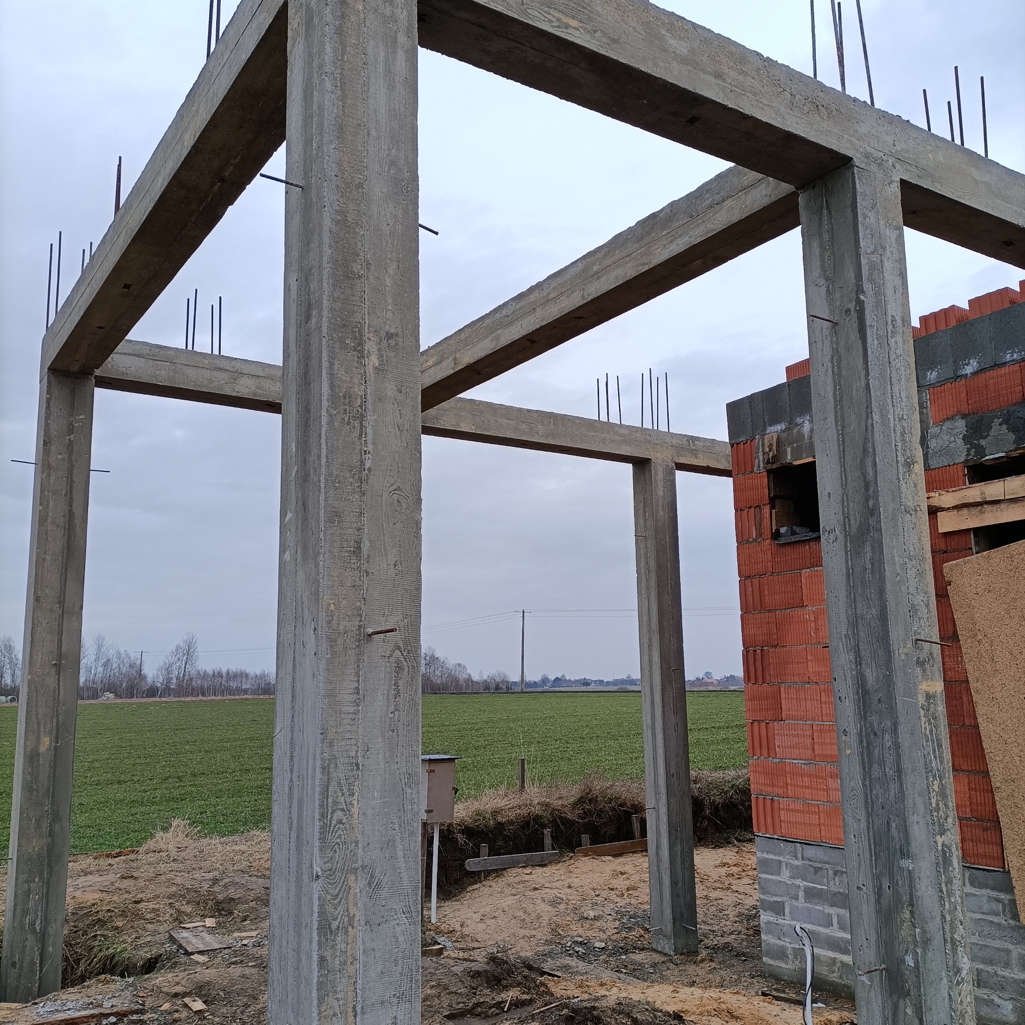 Surowy szkielet domu z betonu z wystającymi prętami zbrojeniowymi, częściowo wzniesione ściany z czerwonej cegły kratówki, widok na pole i pochmurne niebo w tle.