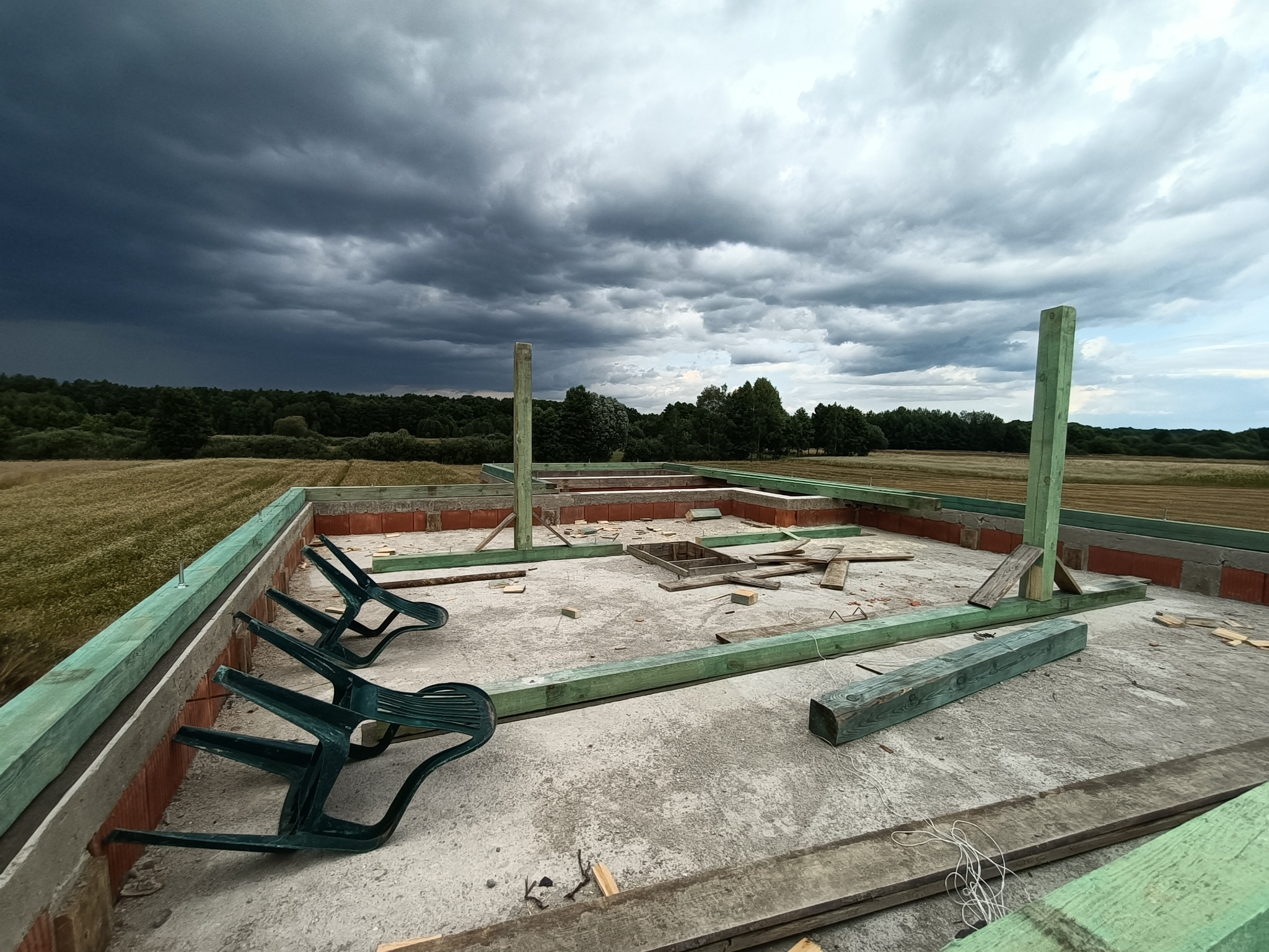 Surowy stan fundamentów domu z cegły i betonu, z zielonymi belkami konstrukcyjnymi i trzema plastikowymi krzesłami na krawędzi, na tle burzowego nieba i pól.