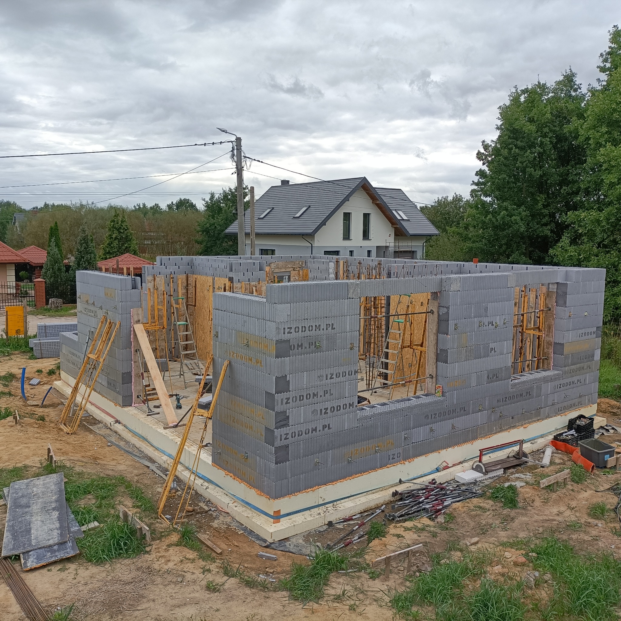 Budowa domu w technologii szalunku traconego: ściany parteru wzniesione z szarych bloczków z widocznymi elementami deskowania i stemplami. W tle widoczny dom w stanie surowym zamkniętym.