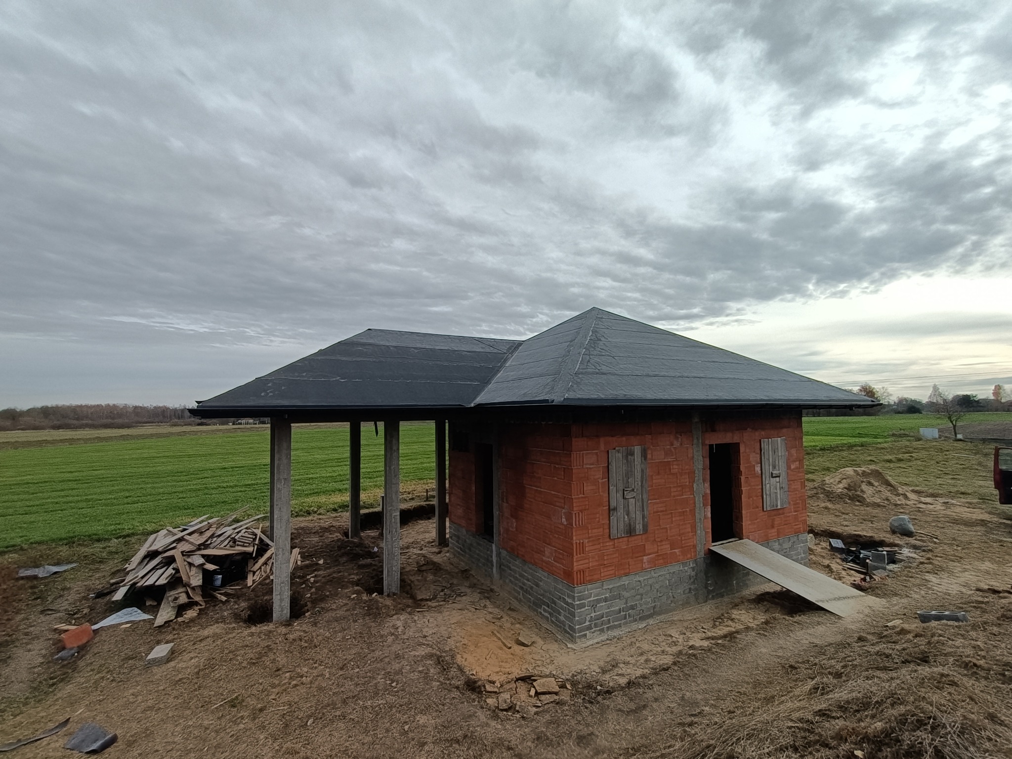 Niedokończony budynek z czerwonej cegły z szarym dachem na tle pola i pochmurnego nieba, widoczne drewniane podpory i prowizoryczna rampa wejściowa.