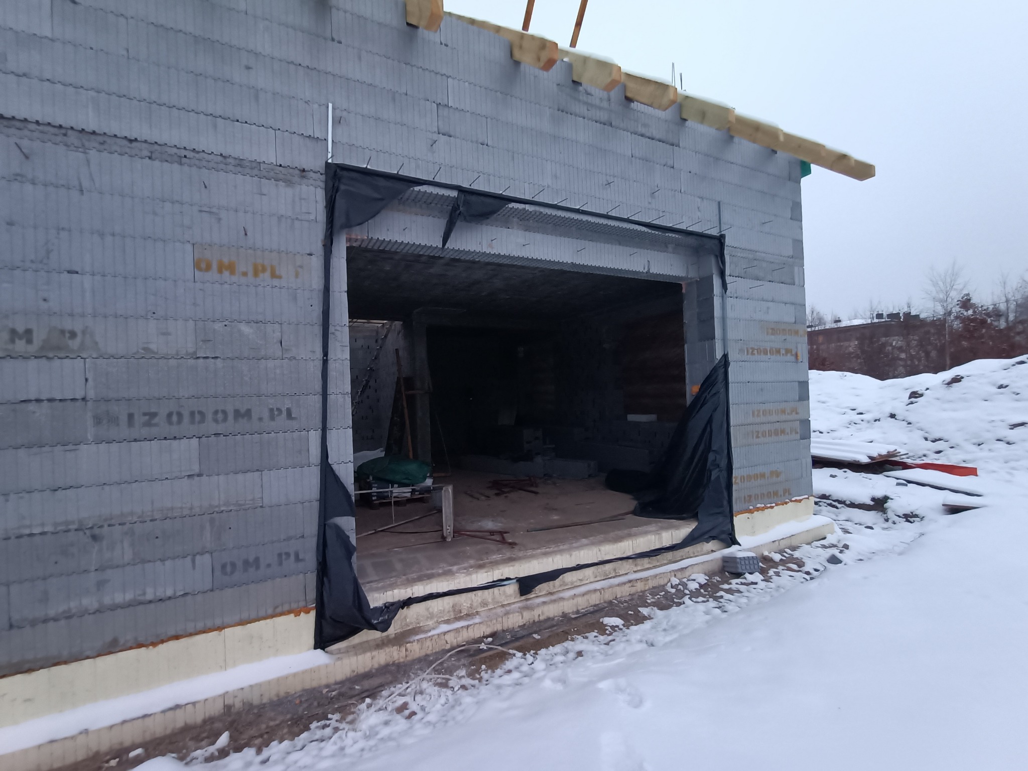 Surowy budynek w trakcie budowy w zimowej scenerii, ściany z szarych bloczków z widocznym logotypem producenta, osłonięte wejście do garażu czarną folią, śnieg na zewnątrz.