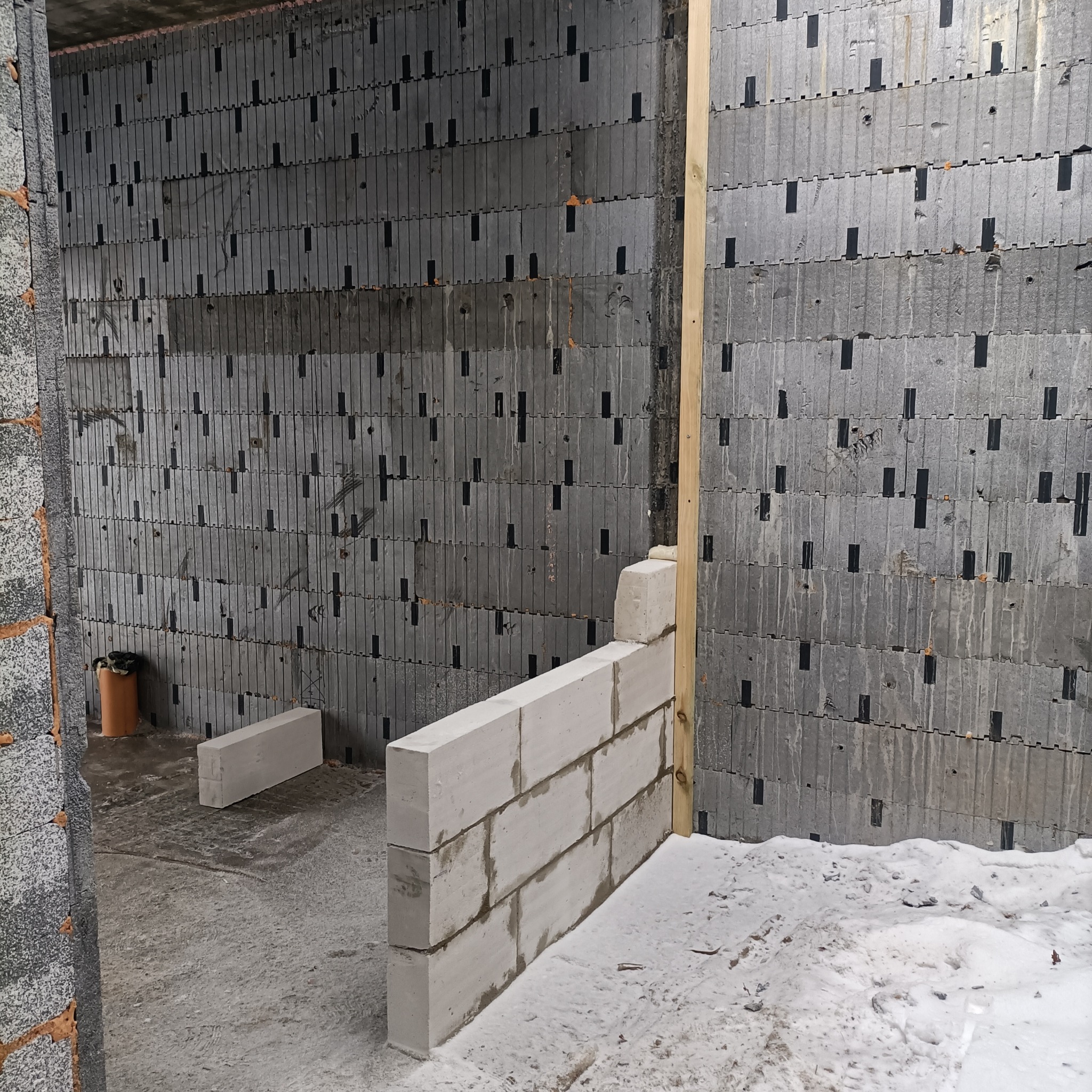 Wnętrze budynku w Nowej Wsi w trakcie budowy, ściany z szarego betonu komórkowego z czarnymi łącznikami, fragment ściany z białych bloczków, drewniana listwa, betonowa podłoga.