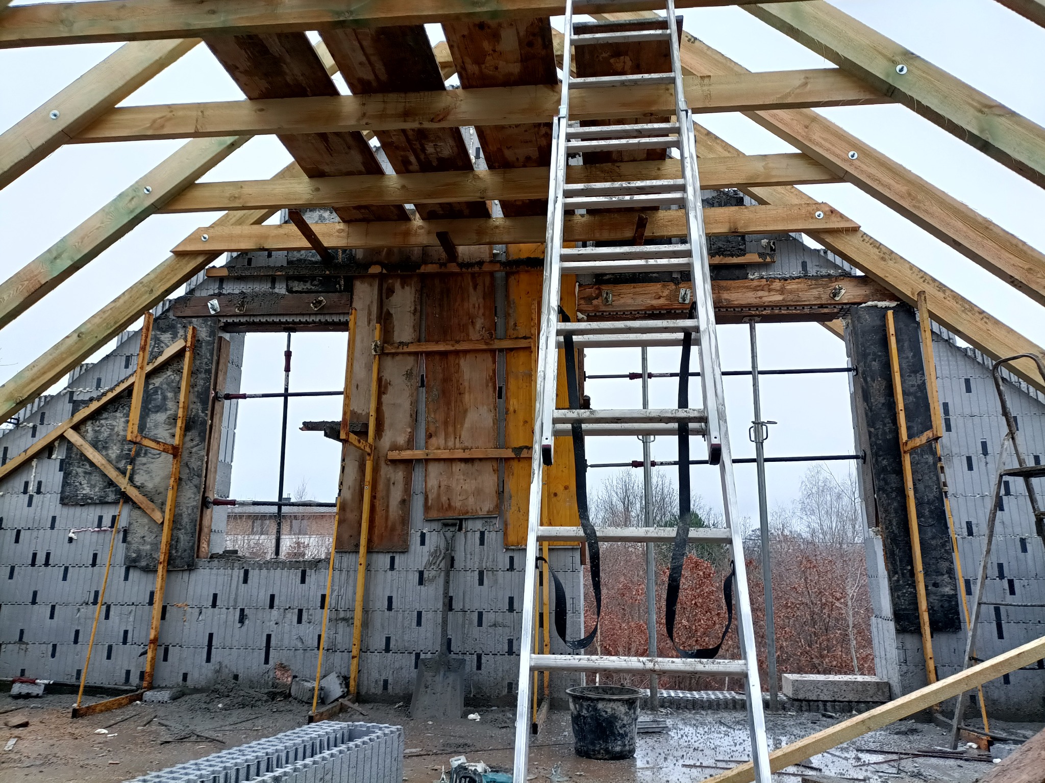 Budowa domu: Ściany z szarego bloczka z widoczną strukturą, drewniana konstrukcja dachu z jasnych belek, aluminiowa drabina oparta o ścianę, widok z wnętrza budynku na zewnątrz przez otwory okienne.