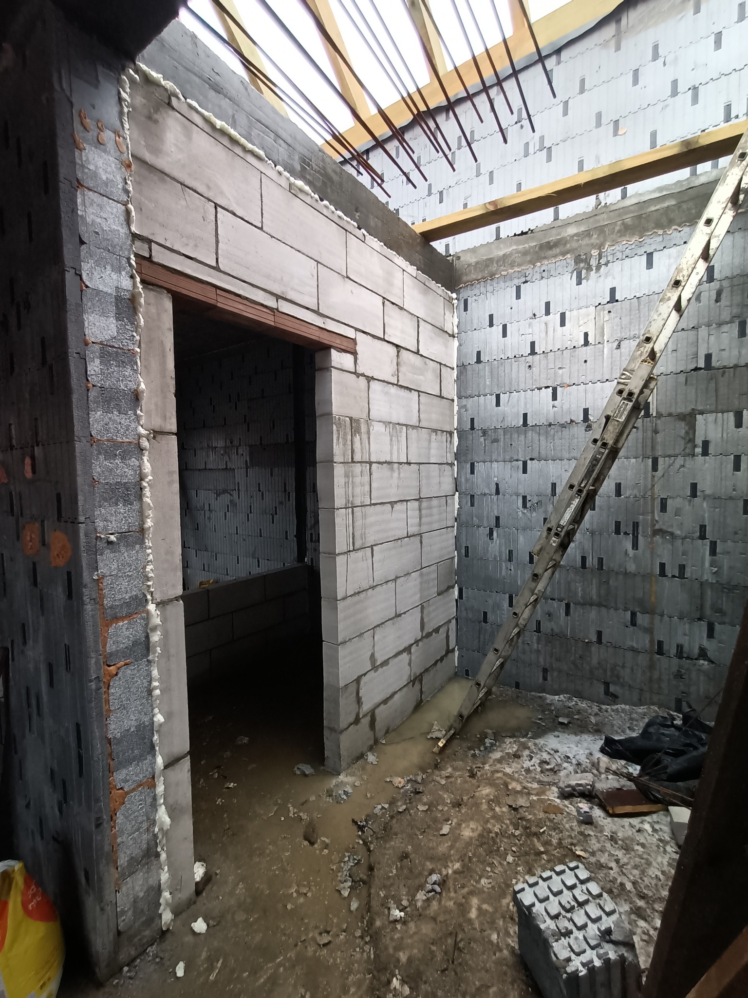 Surowy stan wnętrza budowanego domu z bloków, widoczne ściany z betonu komórkowego, drewniany szkielet dachu, drabina oparta o ścianę, pianka montażowa uszczelniająca ościeżnicę, oraz widoczne...