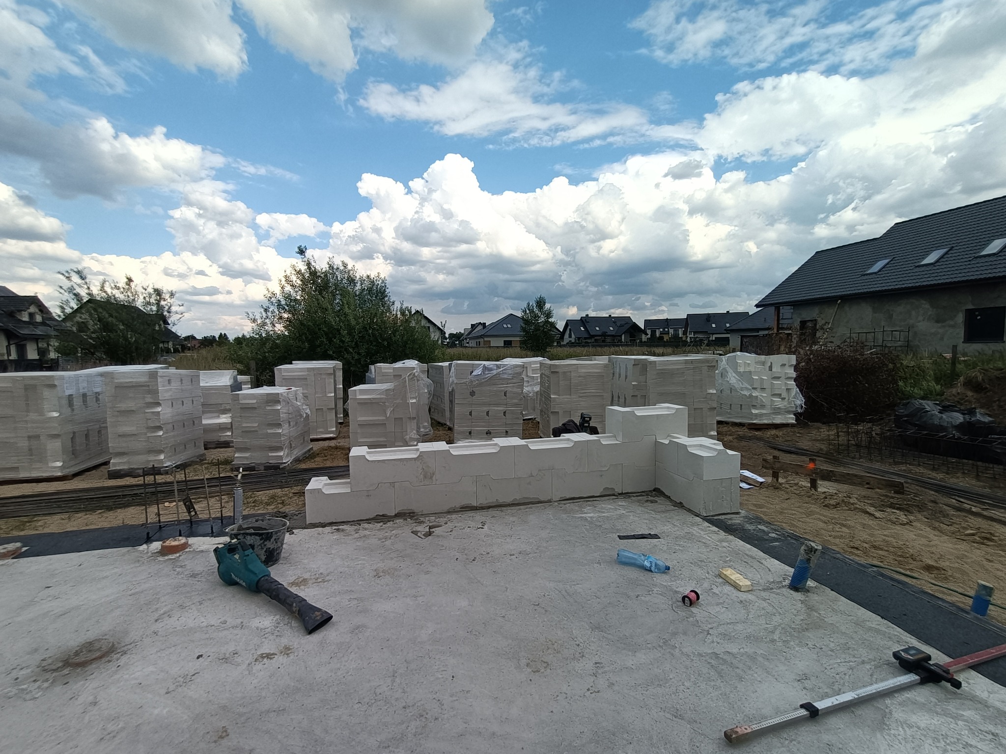 Budowa domu z keramzytu: fundament z częściowo wzniesionymi ścianami, palety bloczków w folii w tle, narzędzia budowlane widoczne na płycie fundamentowej.