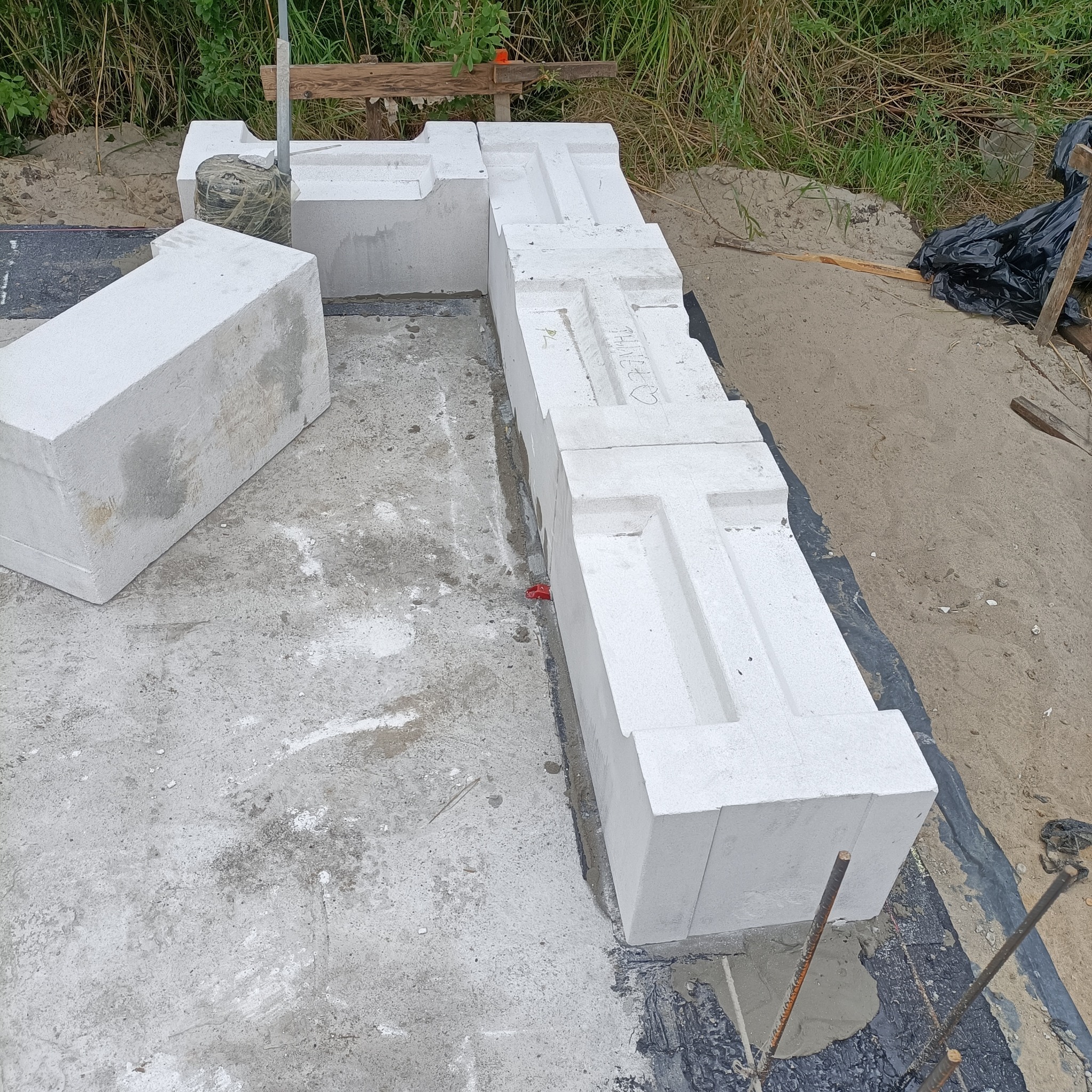 Wznoszenie fundamentów z białych bloczków betonowych o charakterystycznym kształcie T na betonowej płycie, widoczne pręty zbrojeniowe i folia izolacyjna.