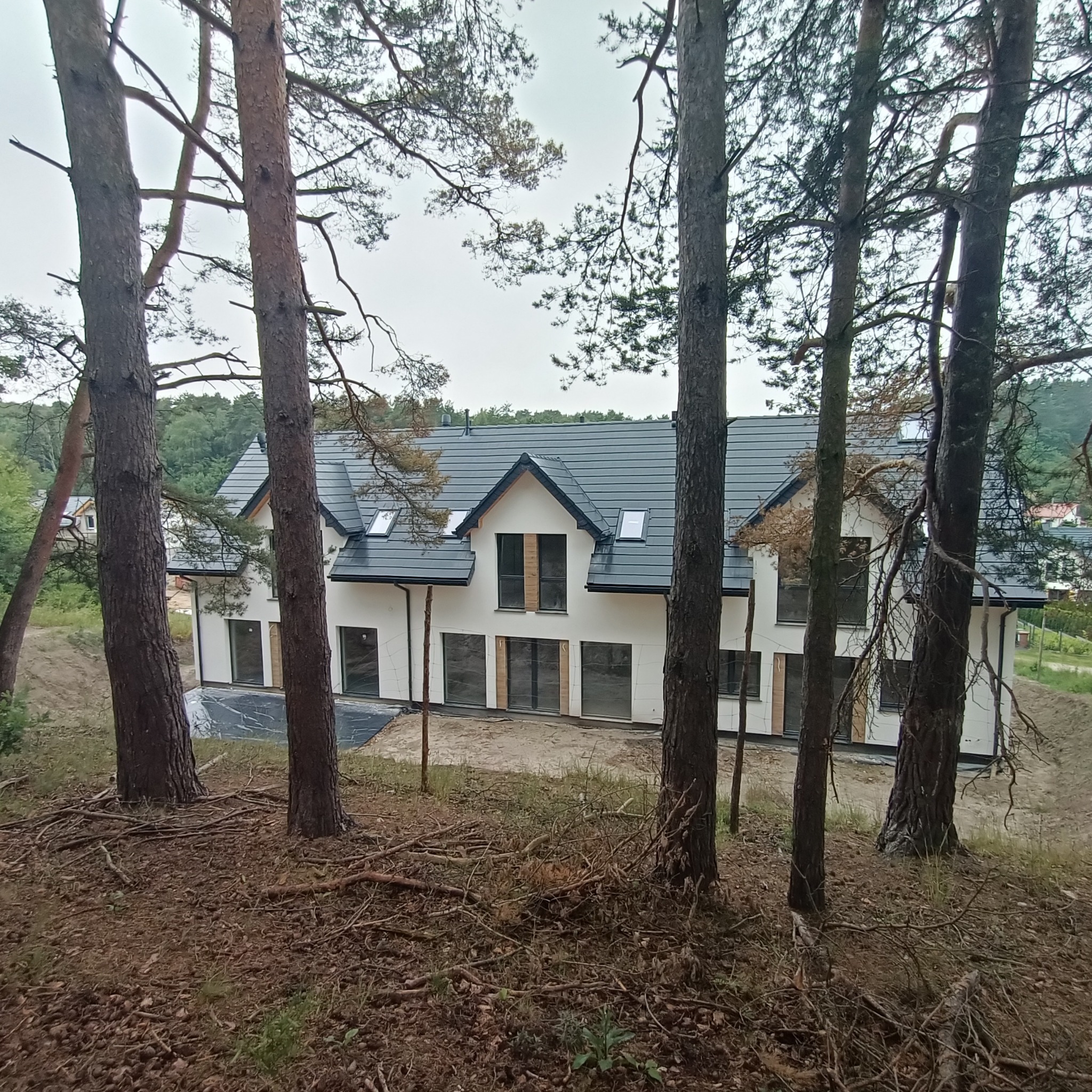 Nowoczesny, trzypiętrowy budynek z białymi ścianami i ciemną dachówką, częściowo zasłonięty przez pnie drzew w lesie. Widoczne okna z drewnianymi elementami dekoracyjnymi. Teren wokół budynku...