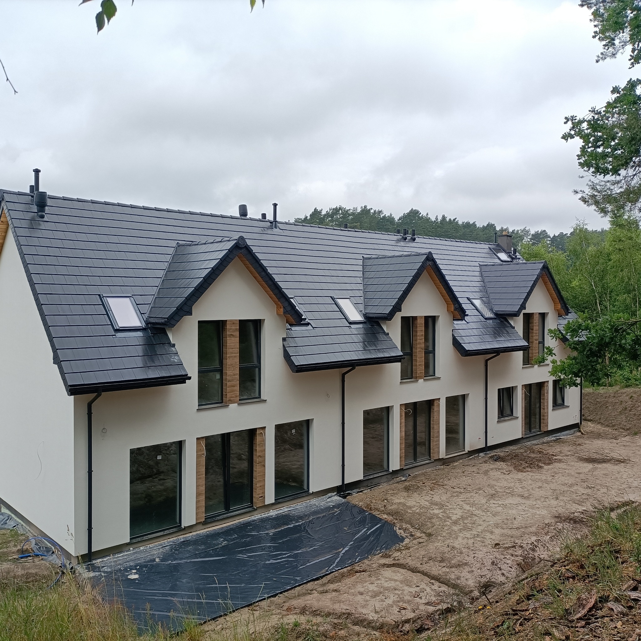 Nowoczesny szeregowiec z białymi ścianami, czarną dachówką i oknami z drewnopodobną okleiną, w trakcie prac wykończeniowych terenu.