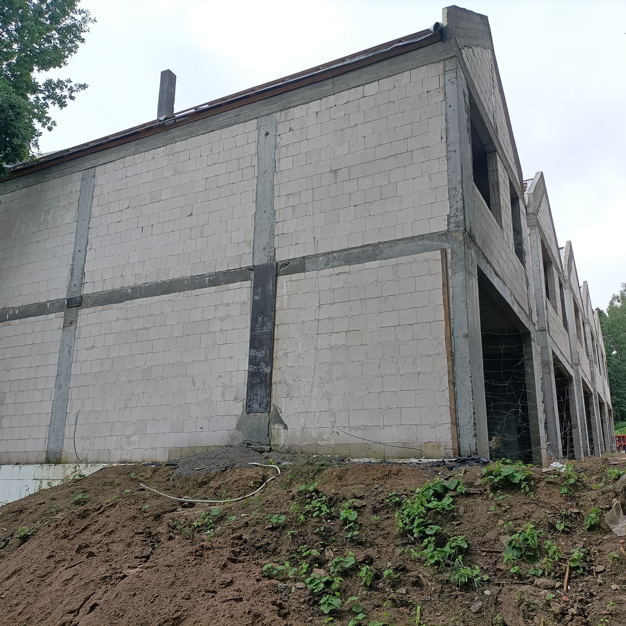 Niedokończony budynek z białych bloczków, widoczne betonowe wzmocnienia konstrukcji, bez okien i drzwi, teren wokół budynku usypany ziemią.