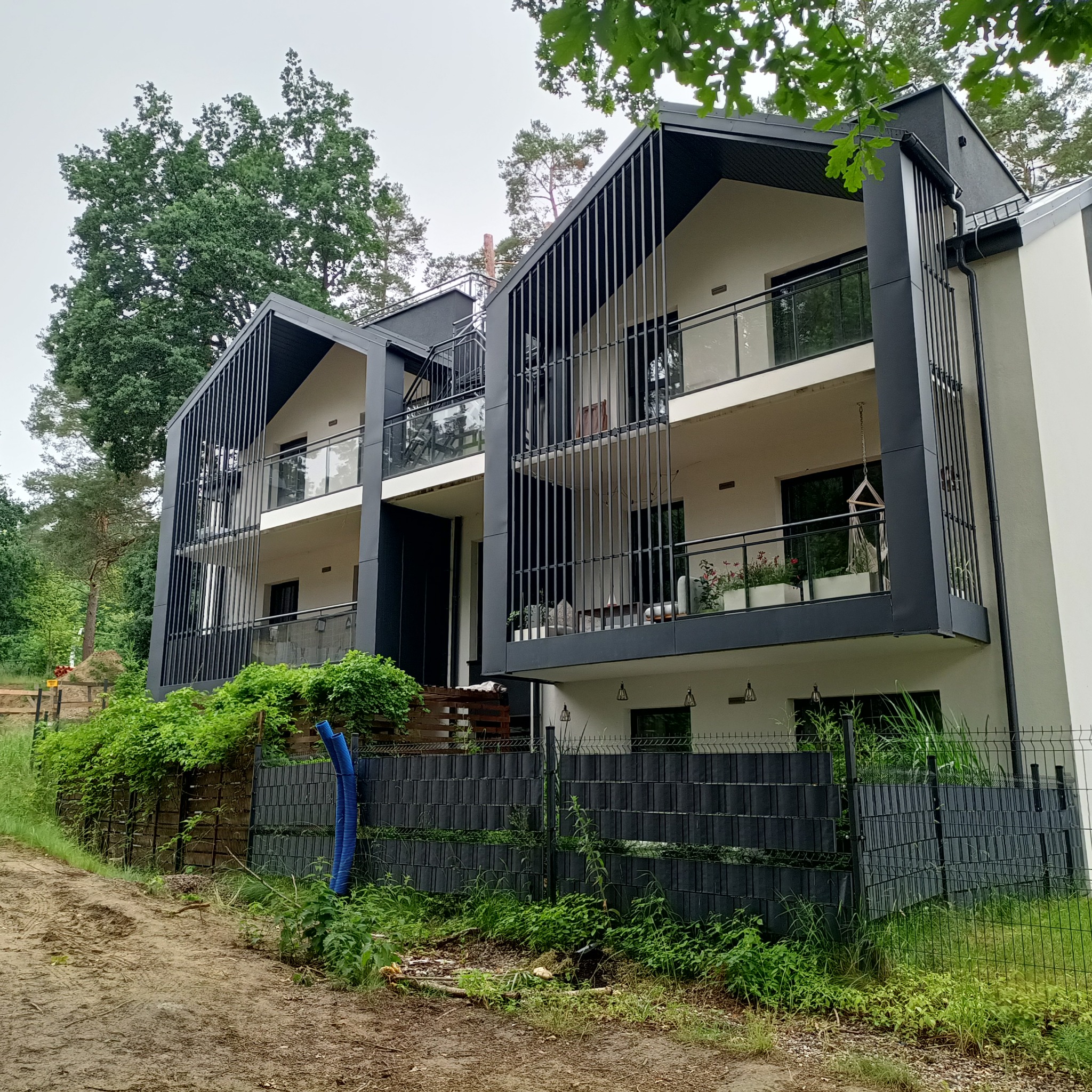 Nowoczesny budynek wielorodzinny z balkonami, wykończony w bieli i czerni, z pionowymi panelami dekoracyjnymi, otoczony zielenią i ogrodzeniem panelowym.