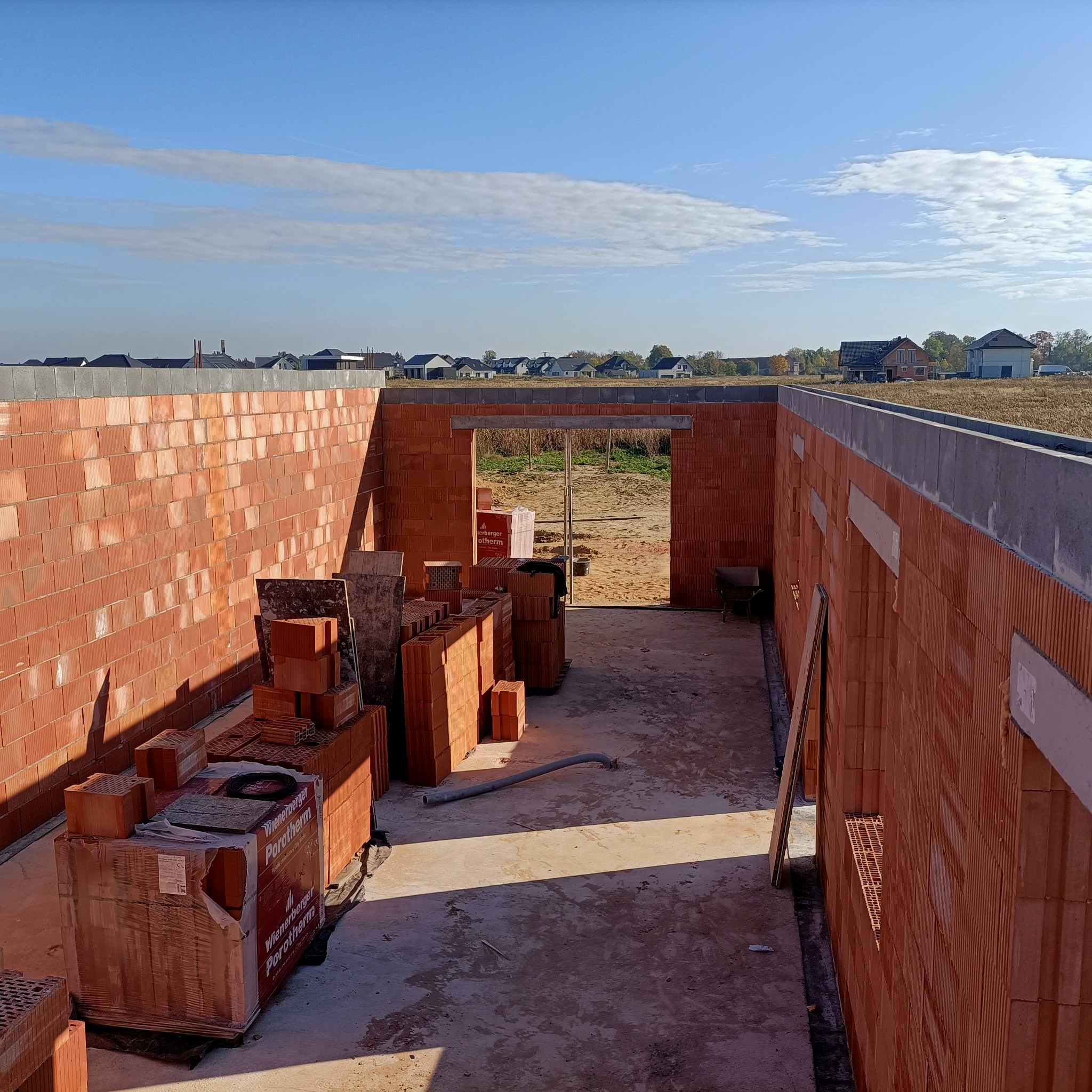 Wnętrze powstającego budynku z czerwonej ceramiki Porotherm, widoczne palety z cegłami, otwory okienne i drzwiowe, w tle zabudowa jednorodzinna pod błękitnym niebem.