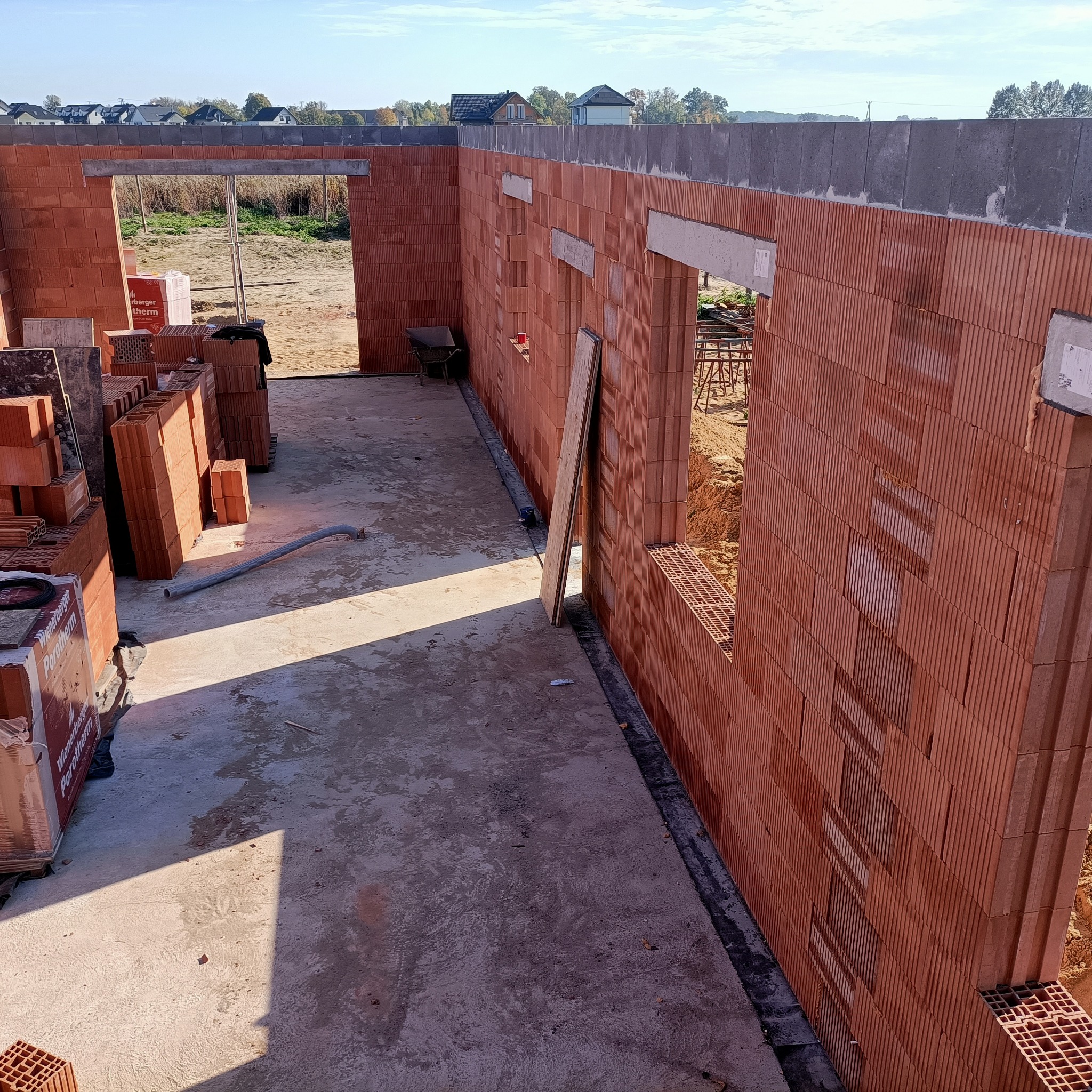 Wnętrze budynku w trakcie budowy z czerwonych pustaków Porotherm, widoczne otwory okienne i drzwiowe, na podłodze leżą materiały budowlane i rura, w tle widać krajobraz.