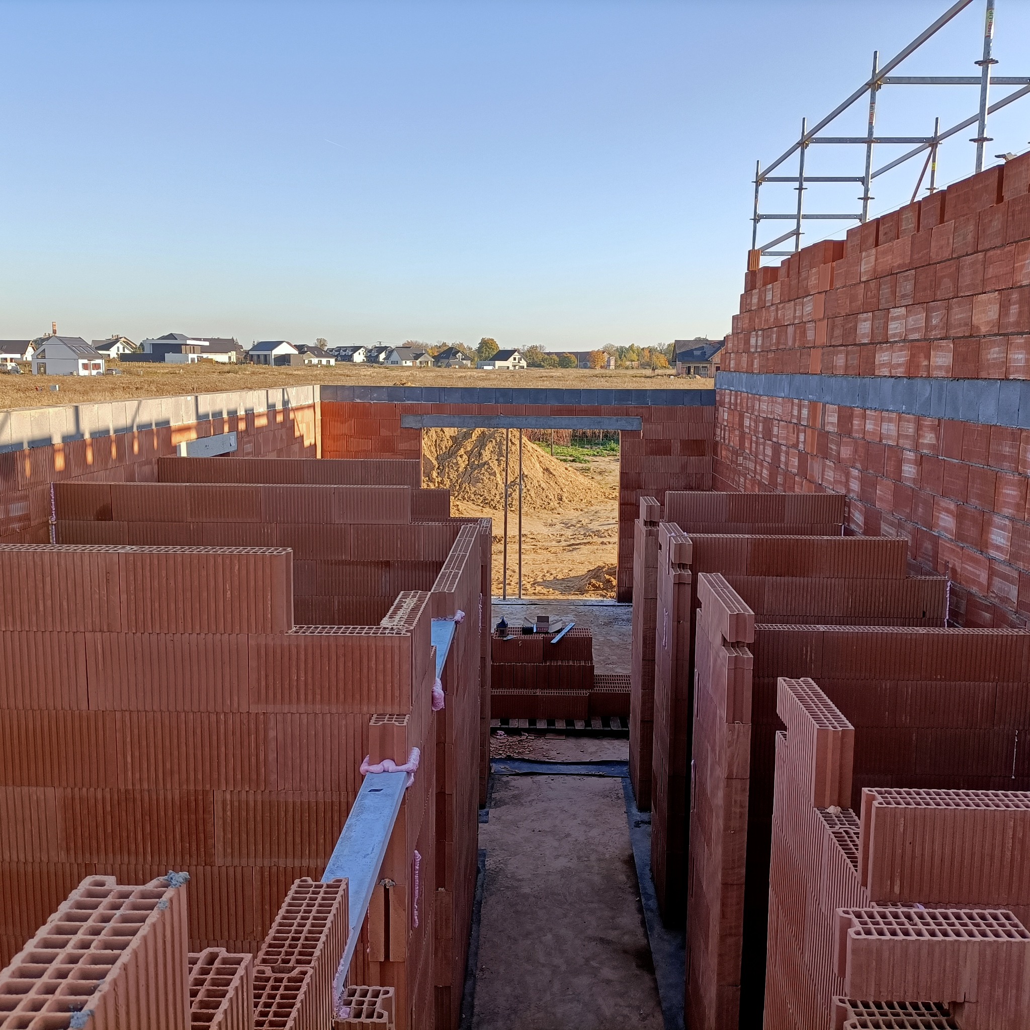 Surowy stan ścian domu z pustaków ceramicznych, widoczne rusztowania i piasek w tle, budowa w trakcie realizacji.