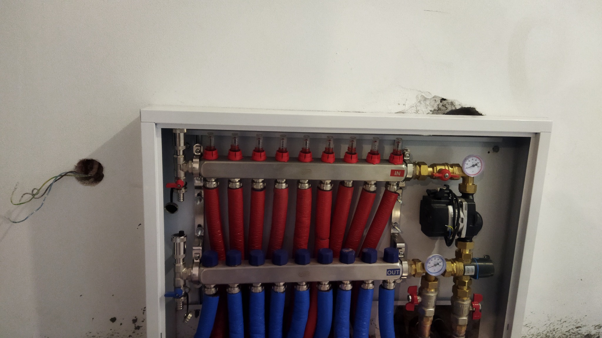 Rozdzielacz ogrzewania podłogowego w szafce, z widocznymi rurami doprowadzającymi i odprowadzającymi wodę, manometrami i pompą obiegową, na tle surowej ściany z wystającymi przewodami elektrycznymi.