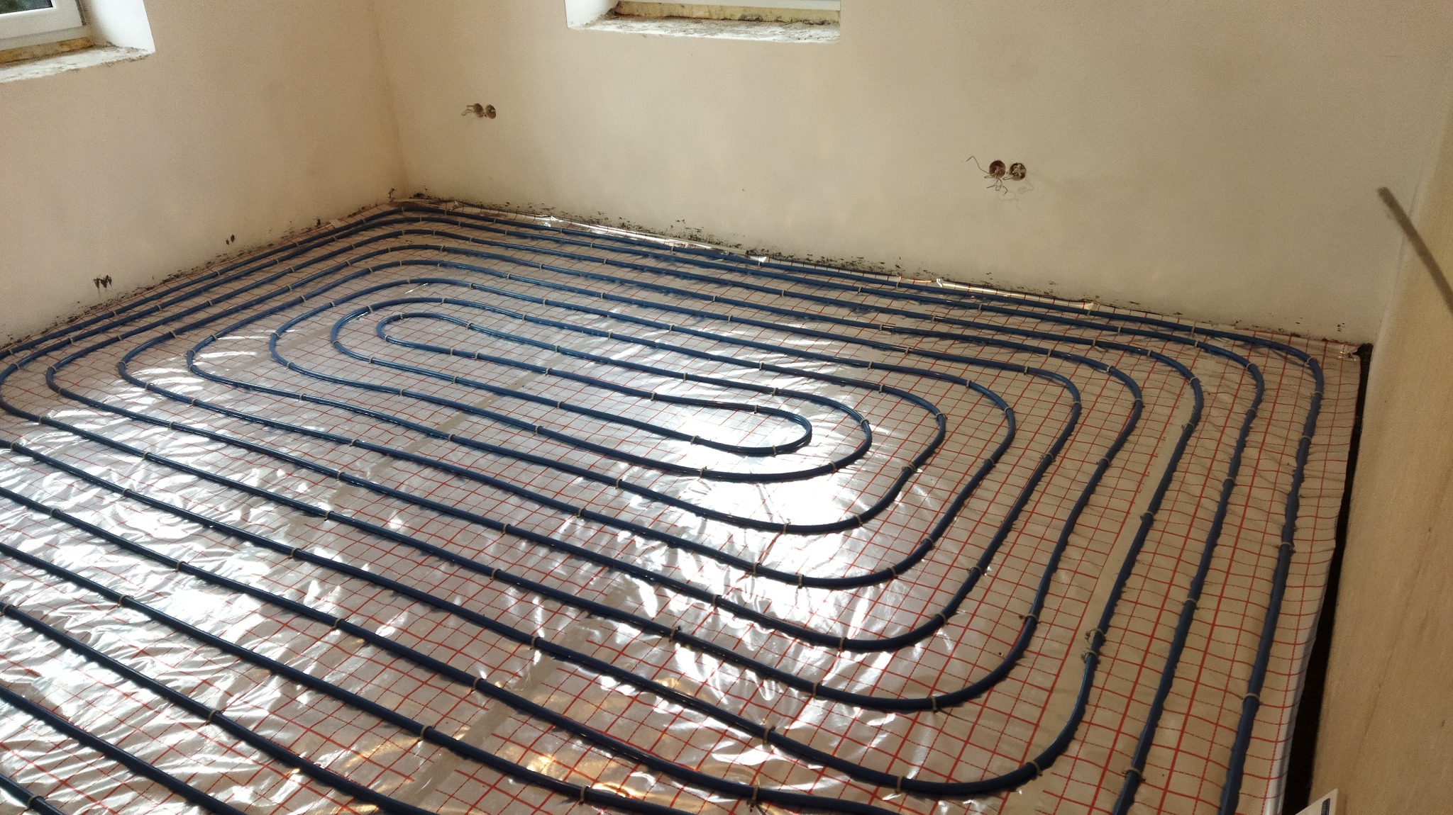 Niedokończony montaż ogrzewania podłogowego z niebieskich rur na folii z czerwoną siatką, widoczne wystające kable elektryczne ze ściany