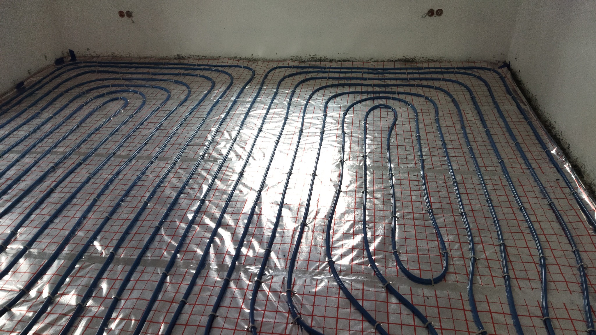 Niebieskie rurki instalacji grzewczej ułożone na podłodze z siatką i folią aluminiową, widok z góry, przygotowanie do wylewki.
