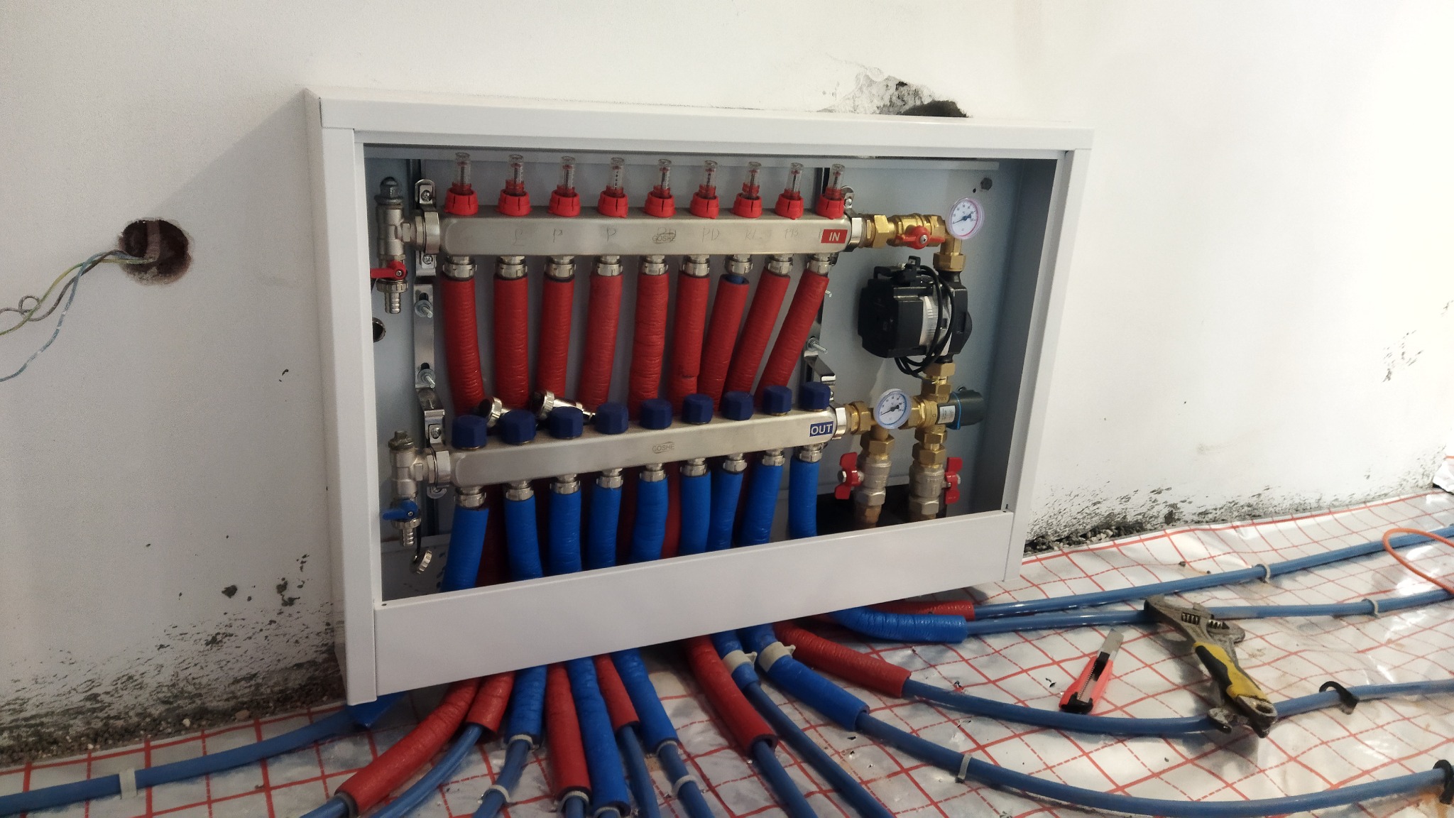 Rozdzielacz ogrzewania podłogowego w białej szafce, z czerwonymi i niebieskimi rurami, widoczny fragment maty grzewczej, klucz nastawny i nóż monterski na podłodze, surowy stan wykończenia ściany.