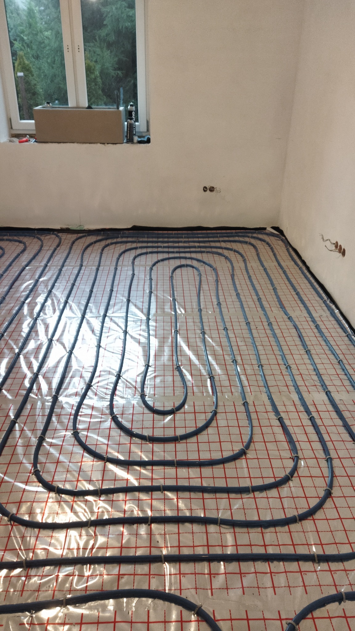 Rozłożone rury ogrzewania podłogowego na czerwonej siatce, widoczne okno i puszki elektryczne w surowym wnętrzu.