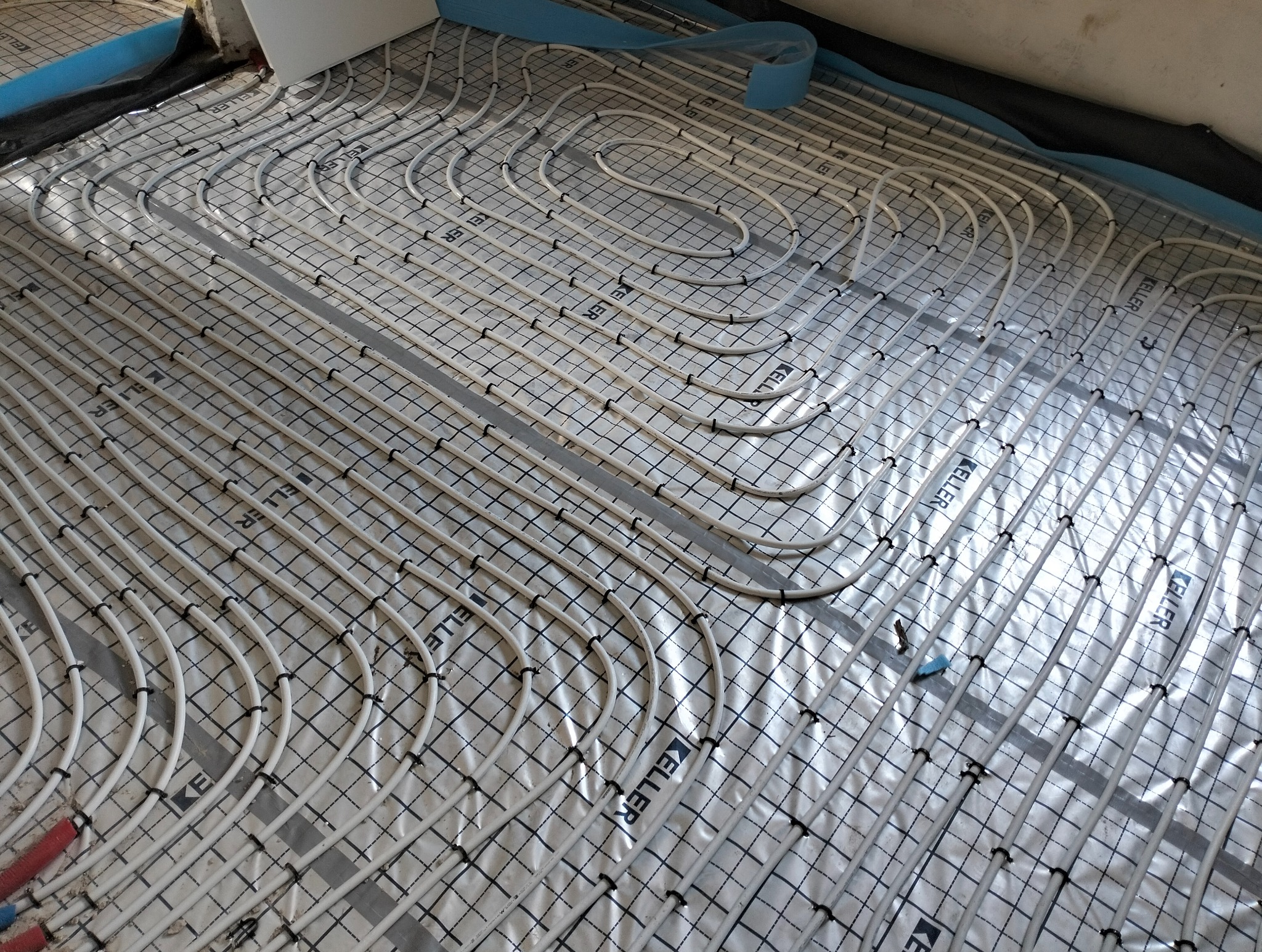 Białe rury ogrzewania podłogowego na srebrnej folii z czarną siatką, zamocowane czarnymi uchwytami. Widoczne niebieskie taśmy i fragment ściany.