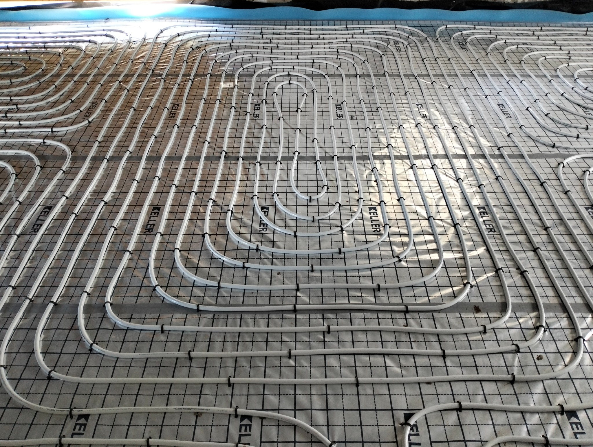 Białe rury ogrzewania podłogowego marki Keller, ułożone w pętle na czarnej siatce z tworzywa, na folii izolacyjnej. Widok z góry, w trakcie montażu.