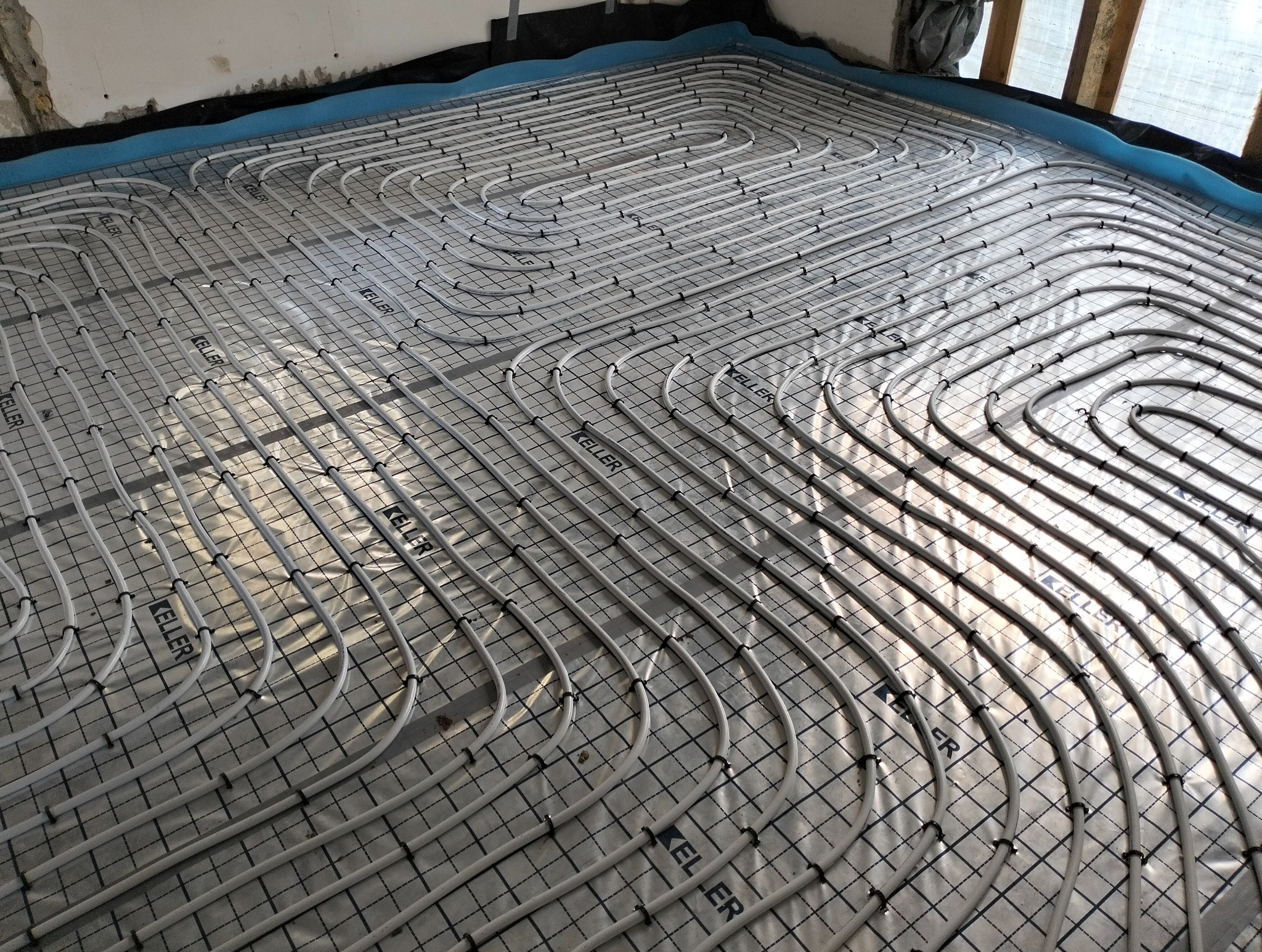 Białe rury ogrzewania podłogowego marki Keller ułożone na folii z rastrem, widok z góry, blisko okna, przed wylaniem posadzki.