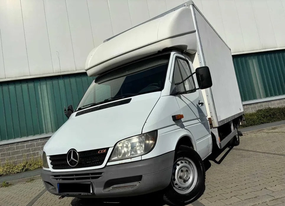 Biały dostawczy Mercedes Sprinter z zabudową kontenerową, widoczny z niskiego kąta, na tle szarej ściany, z lekkim cieniem na karoserii.