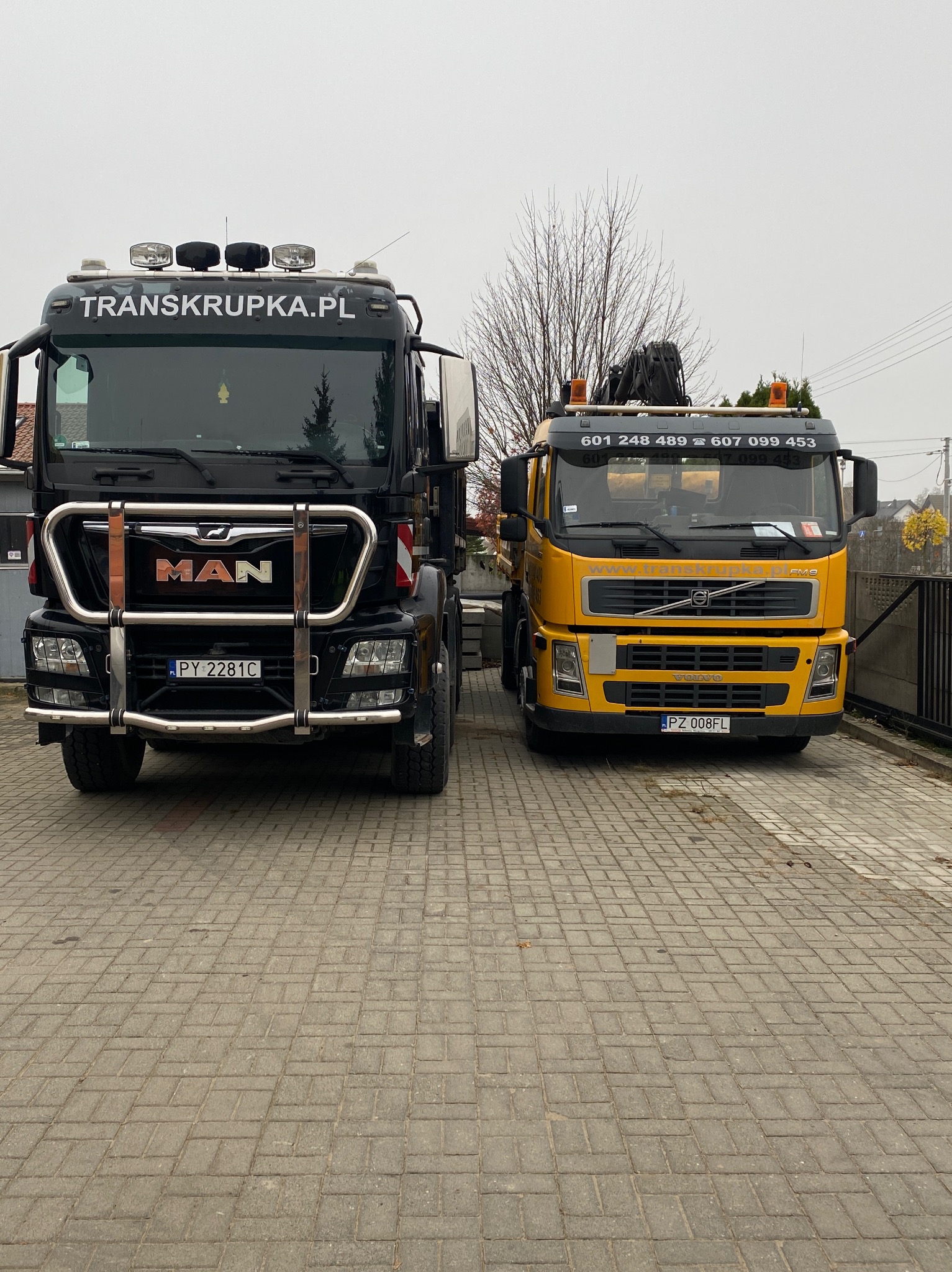 Dwa samochody ciężarowe, czarny MAN z orurowaniem i żółty Volvo z dźwigiem, zaparkowane na kostce brukowej przed ogrodzeniem posesji, z widocznymi numerami telefonów i nazwą firmy TRANSPKRUPKA.PL.