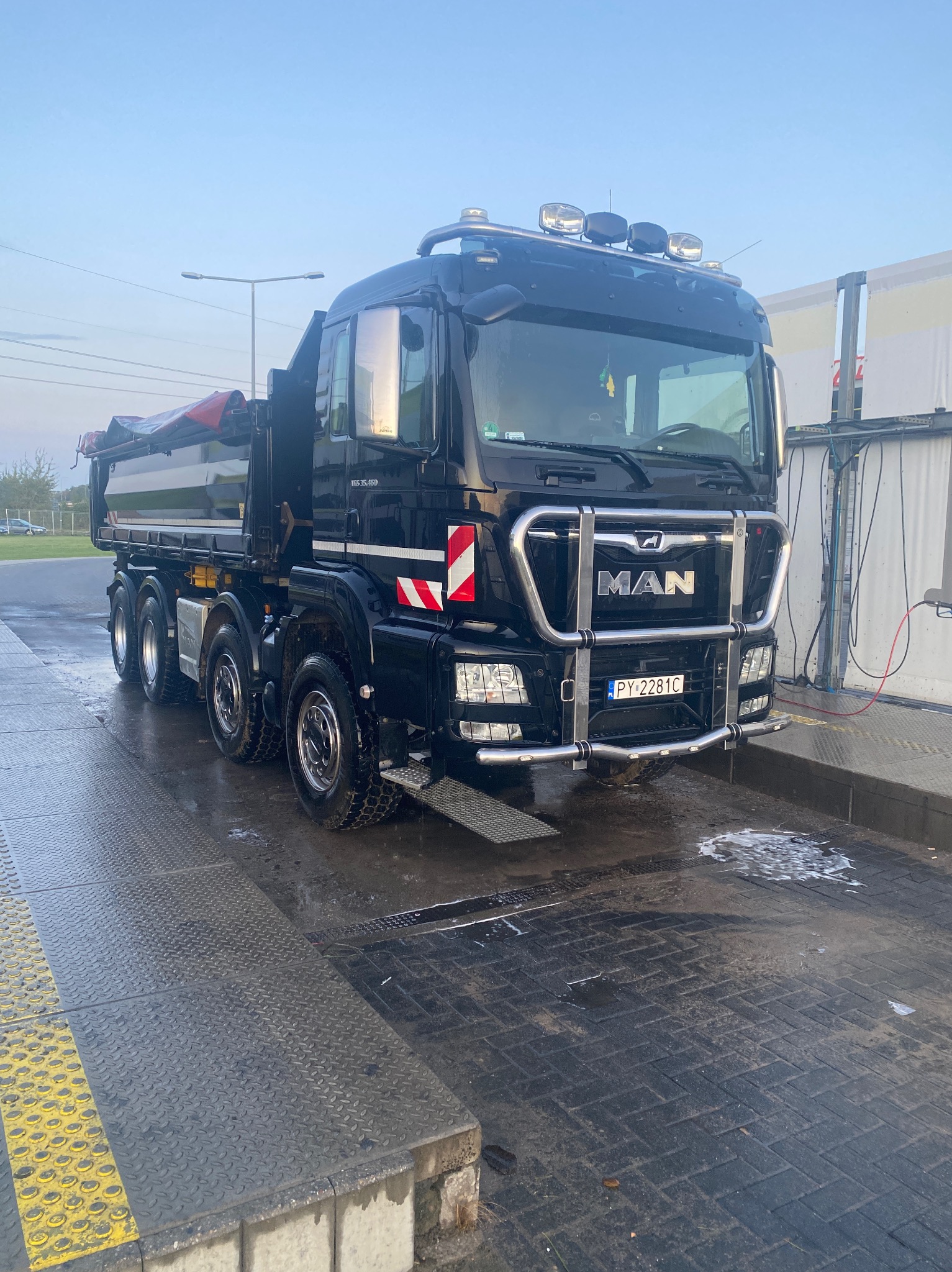 Czarny, błyszczący wywrotka MAN, z widocznymi oznaczeniami ostrzegawczymi, myta w myjni samochodowej, z pianą na posadzce.