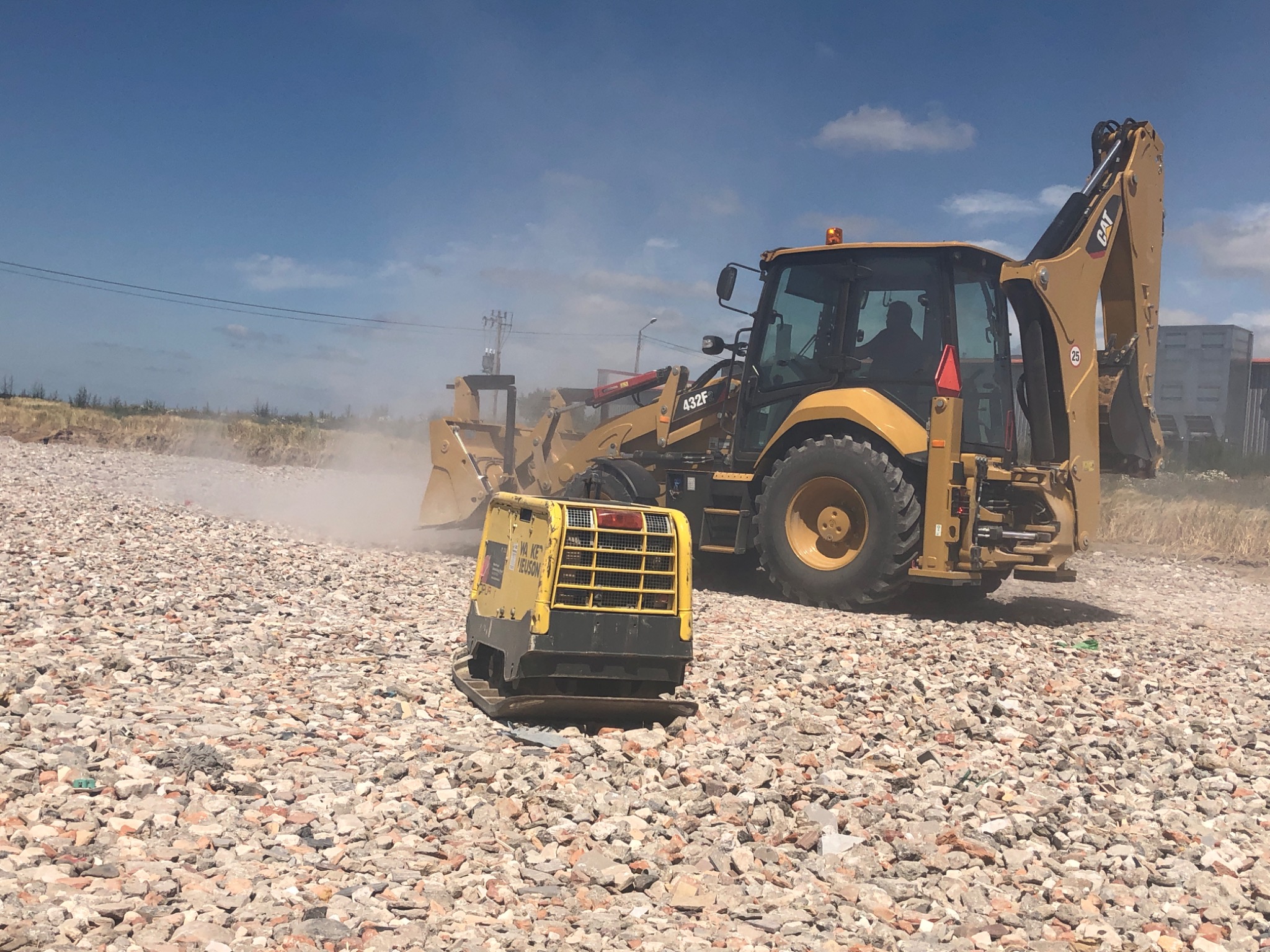 Żółta koparko-ładowarka CAT 432F i zagęszczarka Wacker Neuson na placu budowy, pracujące na obszarze pokrytym gruzem i kamieniami pod błękitnym niebem.