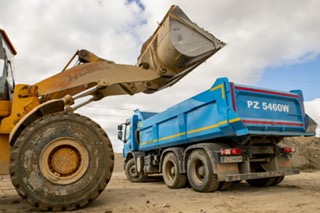 Żółta ładowarka czołowa z pełną łyżką piasku, podczas załadunku na niebieską wywrotkę z otwartą skrzynią, widoczną z tyłu i boku, na placu budowy, w pochmurny dzień.