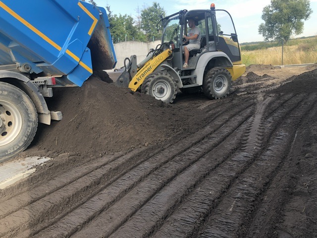 Ciężarówka z niebieską skrzynią wyładowuje ziemię na przygotowany teren, obok stoi żółta ładowarka KramerAllrad z operatorem.