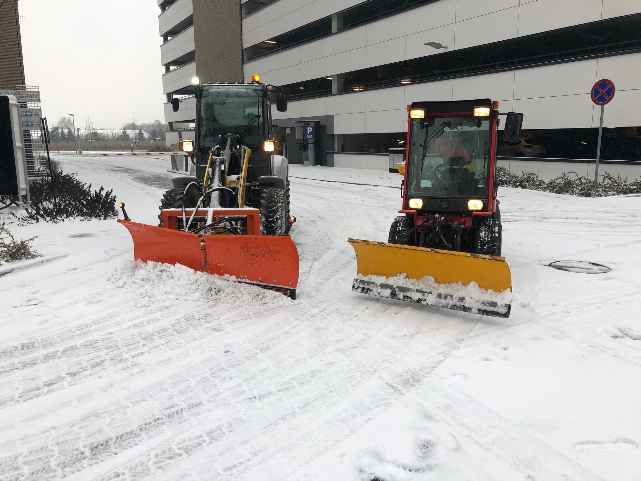 Dwa traktory z pługami, pomarańczowy i żółty, odśnieżają parking wielopoziomowy w zimowy dzień. Widoczne ślady opon w śniegu.