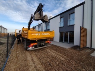 Żółta wywrotka z łyżką koparki na pace, stojąca na gruncie przed nowymi domami w zabudowie szeregowej. Operator w czarnej kurtce przy pojeździe. Widoczny świeżo usypany grunt.