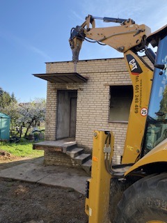 Żółta koparko-ładowarka CAT kruszy młotem pneumatycznym dach murowanego budynku gospodarczego z cegły, widoczne schody wejściowe i brakujące okno, w tle kwitnące drzewa.