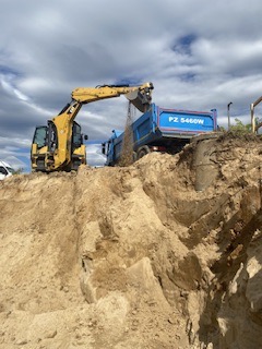 Żółta koparka nabiera piasek z wysokiego nasypu i wsypuje go do niebieskiej ciężarówki z otwartą skrzynią ładunkową, widoczne tablice rejestracyjne. W tle pochmurne niebo.