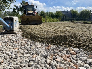 Żwirowo-kamienny podkład pod przyszłą drogę lub plac, z widocznym równiarką i ubijarką w tle, przygotowany pod dalsze prace.