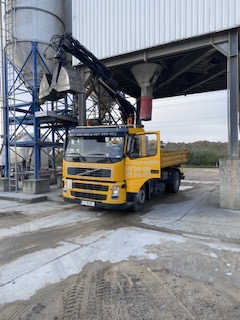 Żółta ciężarówka z dźwigiem i chwytakiem nabiera materiał z silosu. Widoczne ślady opon na ziemi i konstrukcja stalowa nad pojazdem.