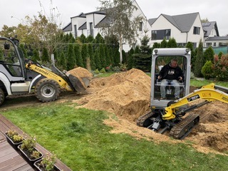 Operator w kabinie mini koparki wykonuje wykop w trawniku, obok ładowarka teleskopowa transportuje ziemię. W tle zabudowa jednorodzinna.