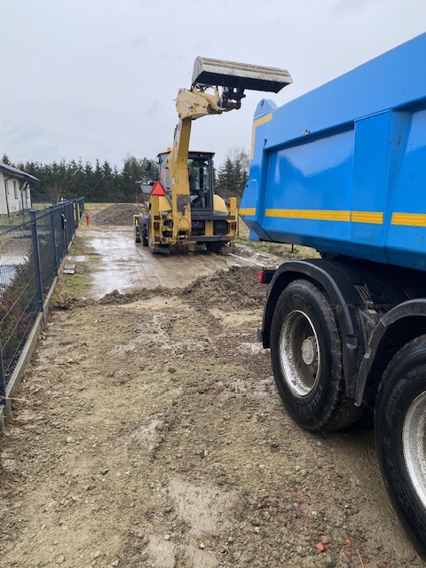 Żółta ładowarka czołowa z uniesioną łyżką wsypuje ziemię do niebieskiej ciężarówki na błotnistym placu budowy, widoczne ogrodzenie posesji i drzewa w tle.