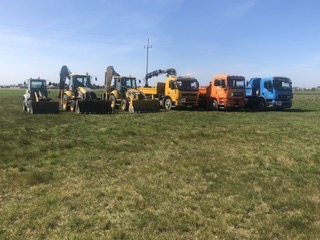 Park maszyn budowlanych: koparki i ciężarówki różnych marek ustawione w rzędzie na trawiastym terenie pod błękitnym niebem.