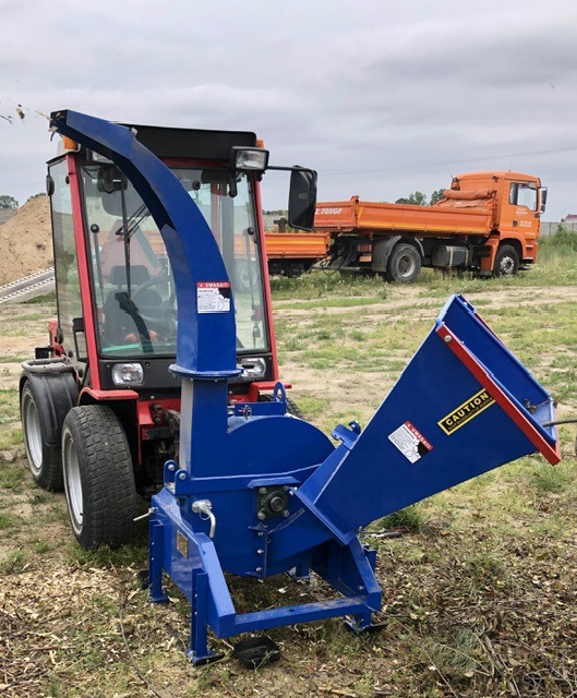 Czerwony ciągnik z niebieskim rębakiem do gałęzi na trawiastym terenie, w tle pomarańczowe wywrotki i usypana ziemia.