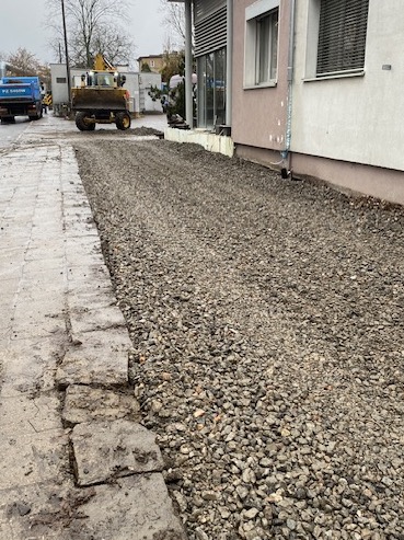 Świeżo wysypany tłuczeń przy budynku, widoczna koparka i ciężarówka w tle, teren przygotowywany do dalszych prac.