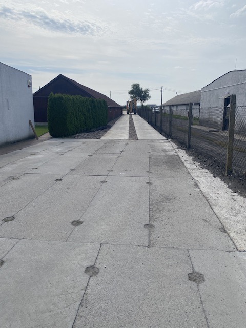 Długa, prosta droga z betonowych płyt z charakterystycznymi okrągłymi wgłębieniami, biegnąca między budynkami i żywopłotem, zakończona koparką w oddali.