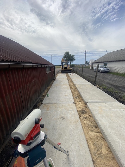 Równanie terenu pod drogę dojazdową z użyciem niwelatora optycznego i koparki; widoczne płyty betonowe tworzące tymczasową nawierzchnię, piaszczyste podłoże i zabudowania gospodarcze w tle.