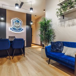 Ministerstwo Korepetycji Łukasz Rudnicki - Nowoczesne wnętrze szkoły korepetycji z podświetlanym logo sowy, biurkiem z laptopem Apple, dwoma niebieskimi krzesłami, wygodną sofą i roślinami doniczkowymi.