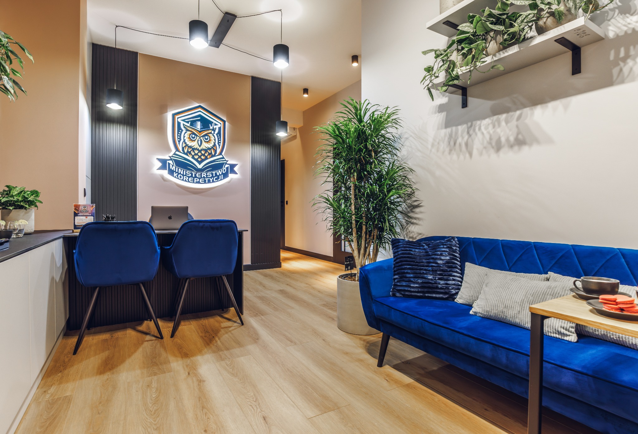 Nowoczesne wnętrze szkoły korepetycji z podświetlanym logo sowy, biurkiem z laptopem Apple, dwoma niebieskimi krzesłami, wygodną sofą i roślinami doniczkowymi.