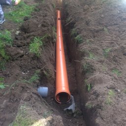 Misza-Bud usługi zimne minikoparką - Pomarańczowa rura kanalizacyjna ułożona w wykopie ziemnym, widoczne ślady świeżo wykopanej ziemi i fragmenty trawy na brzegach wykopu.