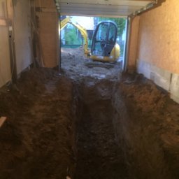 Misza-Bud usługi zimne minikoparką - Wąski wykop w ziemi, wykonany wewnątrz garażu, z widocznym małym żółtym koparką na zewnątrz, oświetlony lampą roboczą wewnątrz.