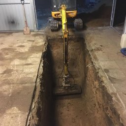 Misza-Bud usługi zimne minikoparką - Żółta mini koparka w wąskim wykopie wewnątrz budynku, obok ściany i łopata oparta o ścianę, słabe oświetlenie.